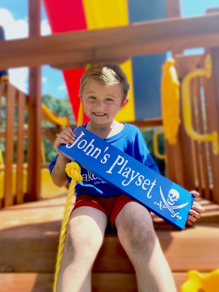 small boy on playset holding up custom sign that reads “John’s playset”